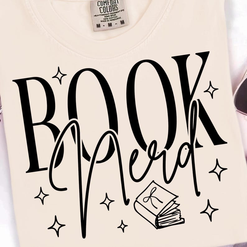 May include: Cream-colored sweatshirt with the words "BOOK Nerd" in large black letters. The word "Nerd" is in a script font. The design includes star shapes and a small book illustration.