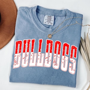 May include: A blue Comfort Colors t-shirt with the word "BULLDOGS" in large, distressed red and white letters. A gold necklace with a pendant rests on the shirt. The shirt is folded on a white surface.