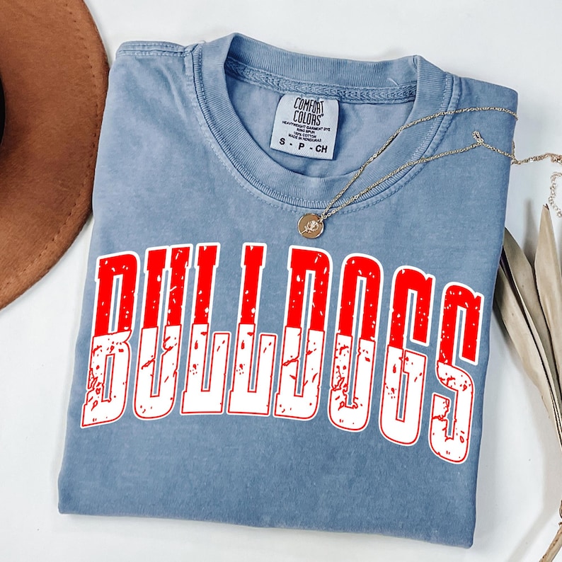 May include: A blue Comfort Colors t-shirt with the word "BULLDOGS" in large, distressed red and white letters. A gold necklace with a pendant rests on the shirt. The shirt is folded on a white surface.