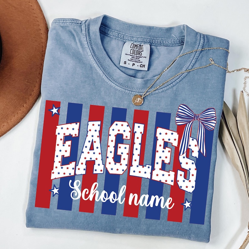 May include: A light blue Comfort Colors t-shirt with the word "EAGLES" in white letters filled with red and blue stars. The shirt has red and blue vertical stripes and a bow graphic. The text "School name" is below the word "EAGLES". A gold necklace is on the shirt.