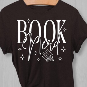 May include: A dark brown t-shirt with the words "BOOK Nerd" in white script and block letters. The design includes a small book illustration and star shapes. The shirt is hanging on a wooden hanger.