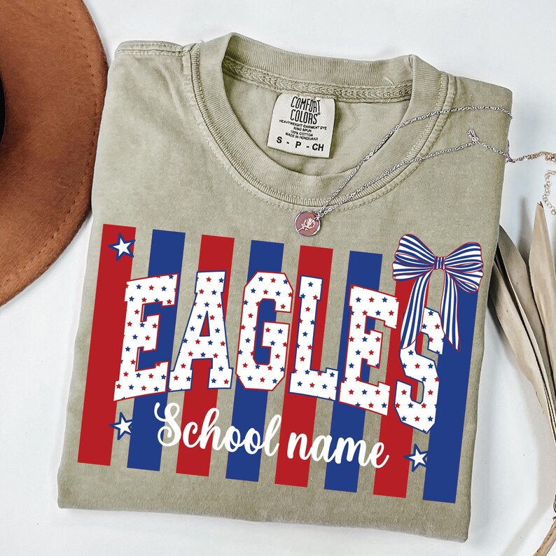 May include: A light olive green t-shirt with a patriotic design. The design features the word "EAGLES" in white block letters filled with red and blue stars, with red and blue stripes. A bow and the words "School name" are also included.