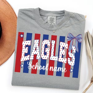 May include: A gray t-shirt with the word "EAGLES" in white letters filled with red and blue stars. Red and blue stripes form a background. The text "School name" is below the word "EAGLES". A bow graphic is on the right side.