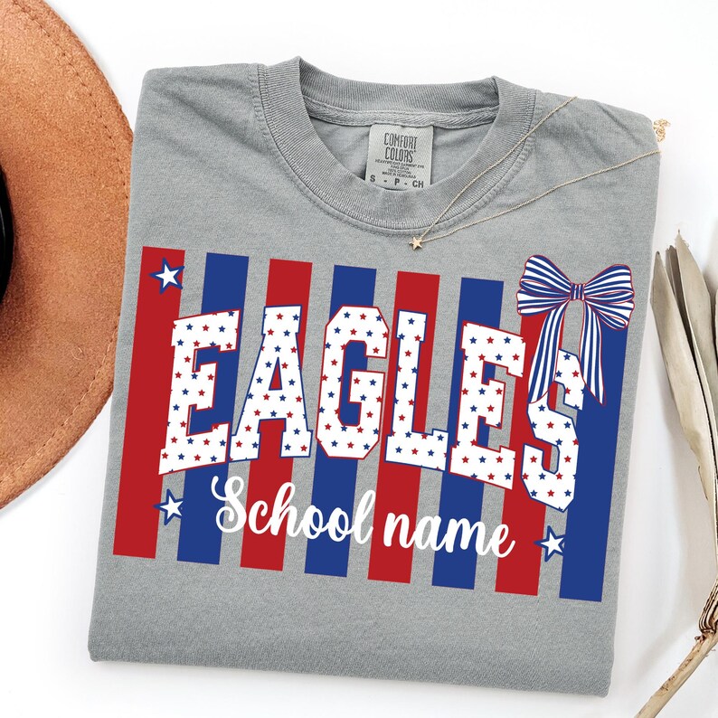 May include: A gray t-shirt with the word "EAGLES" in white letters filled with red and blue stars. Red and blue stripes form a background. The text "School name" is below the word "EAGLES". A bow graphic is on the right side.