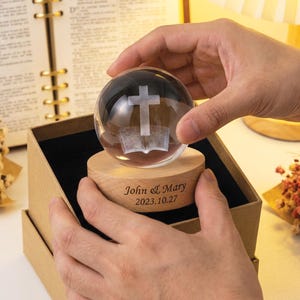 May include: A clear glass sphere with a cross and book design, resting on a wooden base inside a box. The base is engraved with "John & Mary 2023.10.27". The sphere is held by hands.