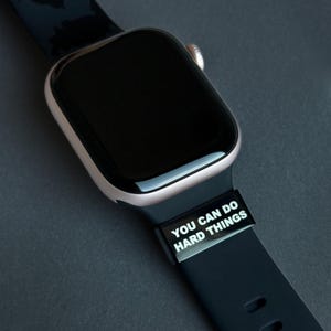 May include: A smartwatch with a black screen and a rose gold case. The black strap features the white text "YOU CAN DO HARD THINGS". The watch is displayed on a dark grey surface.