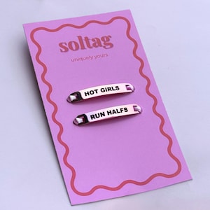 May include: Two metallic barrettes with the phrases "HOT GIRLS" and "RUN HALFS" in black text, displayed on a pink card with the brand name "soltag" and the tagline "uniquely yours". The card has a wavy pink border.