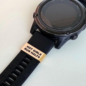 Può includere: Uno smartwatch Garmin nero con cinturino in gomma nero. Una fascia in metallo color oro con la scritta "HOT GIRLS RUN HALFS" è attaccata al cinturino. Il quadrante dell'orologio è nero con dettagli argentati.