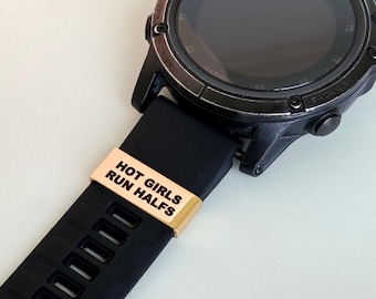 Puede incluir: Un reloj inteligente Garmin negro con una correa de goma negra. Una banda de metal dorada con el texto "HOT GIRLS RUN HALFS" está unida a la correa. La esfera del reloj es negra con detalles plateados.