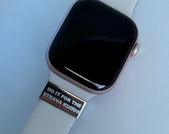 Puede incluir: Un reloj inteligente de oro rosa con una correa blanca. Un accesorio de correa plateada dice "DO IT FOR THE STRAVA KUDOS". La esfera del reloj es negra y la correa se asegura con una hebilla.