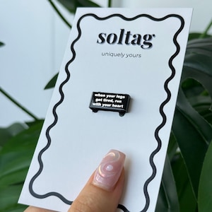 Op de afbeelding: Een zwarte emaille pin met de tekst "when your legs get tired, run with your heart" op een witte kaart met een zwarte golvende rand. De kaart heeft ook de tekst "soltag uniquely yours".