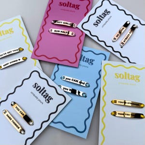 Pode incluir: Uma coleção de etiquetas metálicas com frases motivacionais como "HOT GIRLS" e "YOU'RE STRONGER" exibidas em cartões coloridos. As etiquetas têm um acabamento brilhante e são projetadas para serem presas a roupas ou acessórios.