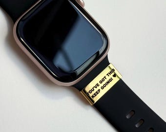 Puede incluir: Un reloj inteligente de oro rosa con una correa negra. Una banda dorada con el texto "YOU'VE GOT THIS KEEP GOING!" está unida a la correa. La esfera del reloj es negra.