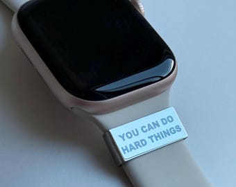 Puede incluir: Primer plano de un reloj inteligente de oro rosa con pantalla negra y correa beige claro. Un pasador de correa plateado con el texto "YOU CAN DO HARD THINGS" está sujeto a la correa.