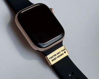 Puede incluir: Un reloj inteligente de oro rosa con una correa negra. Una banda dorada con el texto "YOU'VE GOT THIS KEEP GOING!" está unida a la correa. La esfera del reloj es negra.