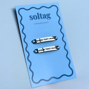 May include: A light blue card with the word "soltag" and the phrase "uniquely yours" printed in white. Two silver-colored name tags with the text "YOUR TEXT HERE" are displayed on the card. The name tags have a blue and white patterned design.