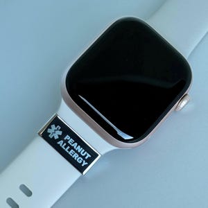 Puede incluir: Un reloj inteligente de oro rosa con pantalla negra y correa blanca. Un dije rectangular negro y plateado en la correa que dice "PEANUT ALLERGY" con un símbolo médico.