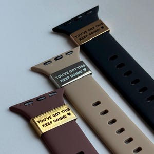 May include: Three Apple Watch straps in black, tan, and brown. Each strap features a metal accent with the text "YOU'VE GOT THIS KEEP GOING!" in white. The straps are made of a flexible material and have adjustment holes.