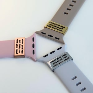 Op de afbeelding: Drie Apple Watch-bandjes in roze, taupe en grijs, elk met een metalen accent gegraveerd met de tekst "when your legs get tired, run with your heart". De bandjes zijn in een stervorm gerangschikt.