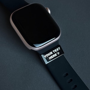 Könnte beinhalten: Nahaufnahme einer Smartwatch mit schwarzem Bildschirm und schwarzem Armband. Auf dem Armband steht in Weiß der Text "YOUR TEXT HERE". Das Uhrengehäuse ist hellgoldfarben.