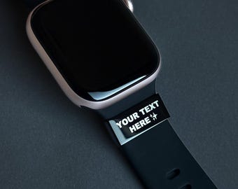 Puede incluir: Primer plano de un reloj inteligente con pantalla negra y correa negra. La correa tiene el texto "YOUR TEXT HERE" en blanco. La caja del reloj es de color oro claro.