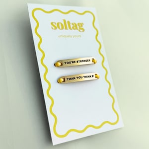 Op de afbeelding: Een witte kaart met een gele rand en de tekst "soltag uniquely yours" in het geel. Twee goudkleurige metalen staven zijn bevestigd, elk met een geel koord en gegraveerde tekst: "YOU'RE STRONGER" en "THAN YOU THINK ❤".