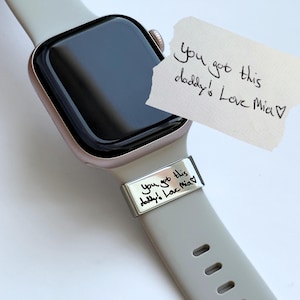 以下が含まれることがあります： ローズゴールドのスマートウォッチとライトグレーのシリコンバンド。バンドには「you got this daddy! Love Mia♡」という手書き文字が刻印された長方形のメタルチャームが付いています。同じ手書きメッセージが書かれた小さな紙が近くに置かれています。
