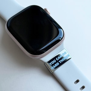 Puede incluir: Primer plano de un reloj inteligente con una correa blanca y un dije plateado. El dije tiene las palabras "YOU CAN DO HARD THINGS" en texto azul. La esfera del reloj es negra.