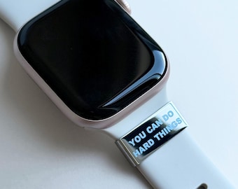 Puede incluir: Primer plano de un reloj inteligente con una correa blanca y un dije plateado. El dije tiene las palabras "YOU CAN DO HARD THINGS" en texto azul. La esfera del reloj es negra.