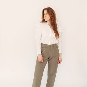 May include: A woman wearing a white button-up shirt with puffed sleeves and olive green pants. She is standing in front of a white background.