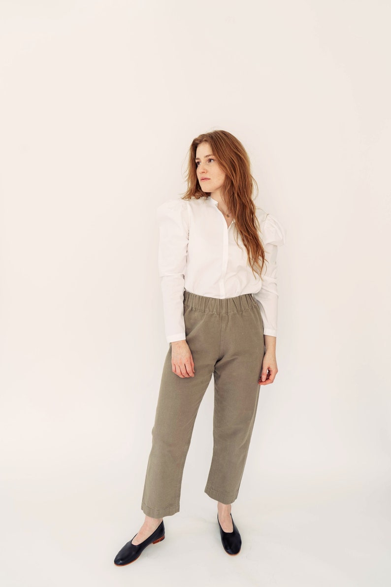 May include: A woman wearing a white button-up shirt with puffed sleeves and olive green pants. She is standing in front of a white background.