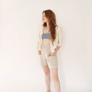 May include: A woman wearing a white cardigan, a blue and white gingham top, beige linen shorts, and brown leather shoes. She is standing in front of a white background.