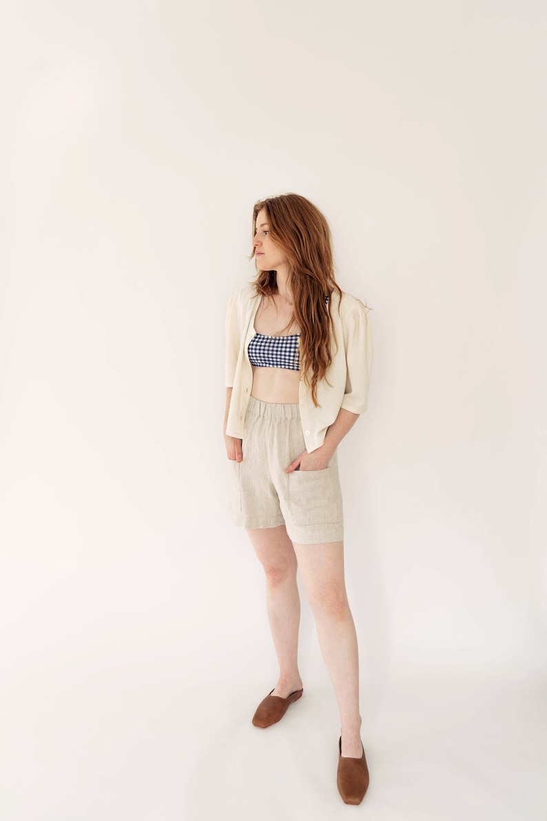 May include: A woman wearing a white cardigan, a blue and white gingham top, beige linen shorts, and brown leather shoes. She is standing in front of a white background.