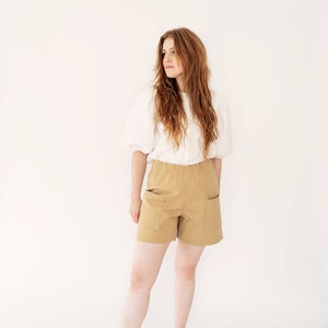 May include: A woman wearing a white blouse and khaki shorts with pockets stands in front of a white background. She is wearing brown leather mules.