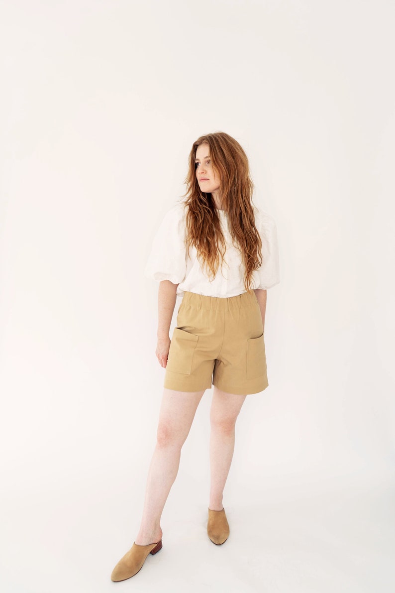 May include: A woman wearing a white blouse and khaki shorts with pockets stands in front of a white background. She is wearing brown leather mules.