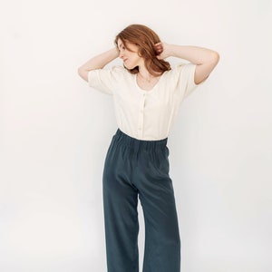 May include: A woman wearing a white, button-up blouse with puffed sleeves and dark green, wide-leg pants. She is standing with her arms raised above her head, looking down.