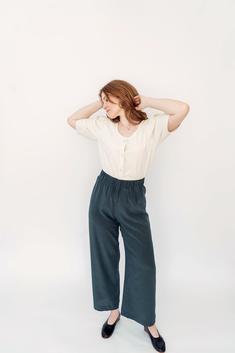 May include: A woman wearing a white, button-up blouse with puffed sleeves and dark green, wide-leg pants. She is standing with her arms raised above her head, looking down.