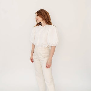 May include: A woman wearing a white, button-up blouse with puffed sleeves and white, straight-leg pants. She is wearing brown leather flats.