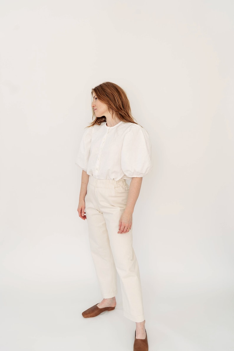 May include: A woman wearing a white, button-up blouse with puffed sleeves and white, straight-leg pants. She is wearing brown leather flats.