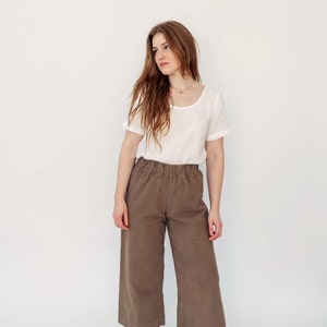 May include: A woman wearing a white linen top and brown wide-leg pants with a relaxed fit. She is wearing brown leather lace-up boots.