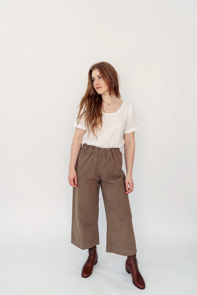 May include: A woman wearing a white linen top and brown wide-leg pants with a relaxed fit. She is wearing brown leather lace-up boots.