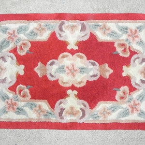 May include: A small red rug with a floral pattern and white fringe. The rug has a central design of stylized flowers and leaves in shades of pink, white, and green.