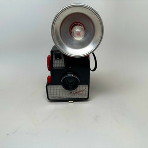 May include: A black and red vintage camera with a silver flash attachment. The camera has a built-in flash and says "Imperial Automatic" on the front.