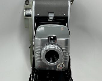 Puede incluir: Cámara Polaroid vintage con lente plateada y cuerpo gris. La cámara tiene una correa de cuero negro y un panel frontal negro que se abre. La palabra "Polaroid" está impresa en la parte delantera.