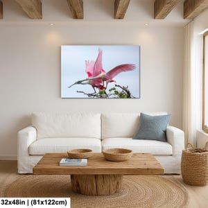 a large, vibrant pink bird in flight, captured in a striking photograph. The bird is the central focus of the image, with its wings spread wide as it soars through the sky. The photograph is displayed on a wall above a white couch, creating a cozy and inviting atmosphere in the living room.