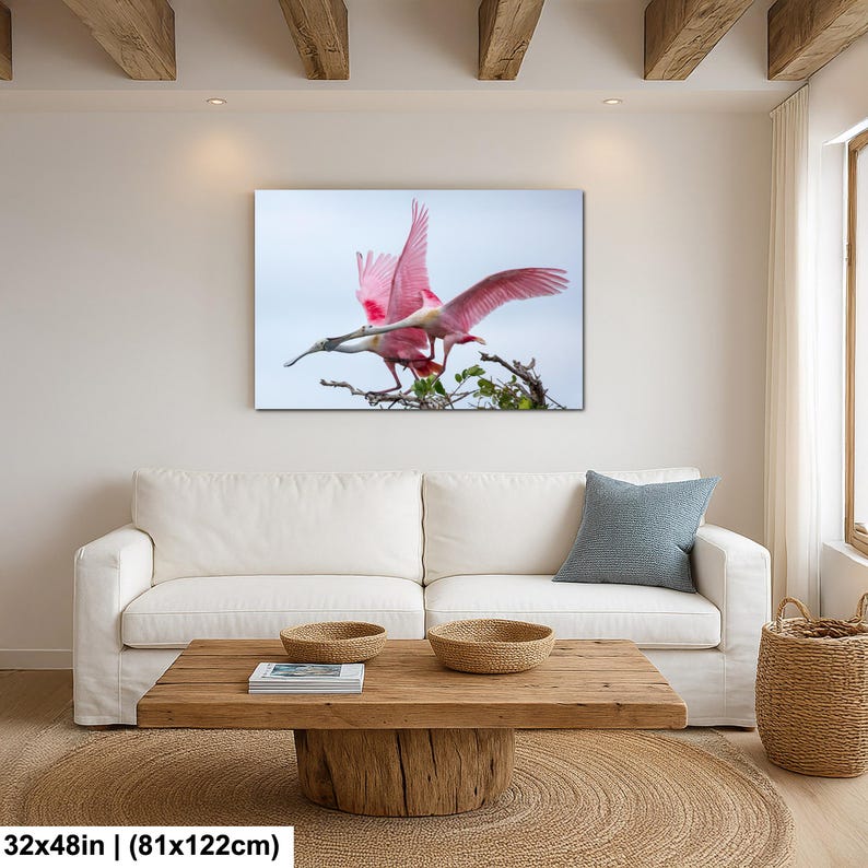 a large, vibrant pink bird in flight, captured in a striking photograph. The bird is the central focus of the image, with its wings spread wide as it soars through the sky. The photograph is displayed on a wall above a white couch, creating a cozy and inviting atmosphere in the living room.