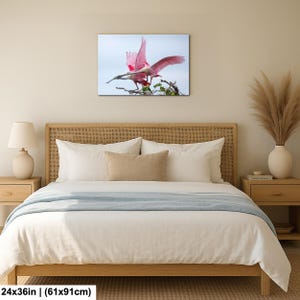 a bedroom with a large bed, a nightstand, and a lamp. Above the bed, there is a framed photograph of a pink bird with outstretched wings.