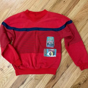 May include: Red striped sweatshirt with a navy blue stripe across the chest. The sweatshirt has two patches on the front, one with the text "The American Discovery Trail" and the other with the text "Follow the Highway" and a Native American design.