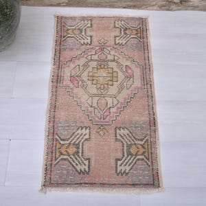 May include: A rectangular, vintage rug with a faded, distressed appearance. The rug features a central geometric design in beige, pink, and gold, surrounded by a pink and gray border. The rug's dimensions are approximately 24 x 48 inches.