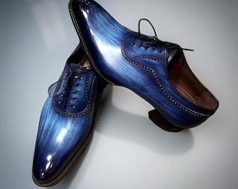 May include: A pair of oxford shoes featuring a polished finish with a gradient blue and black brushstroke pattern. The shoes have a closed lacing system, decorative brogue detailing along the seams, thin black laces, and a dark brown sole.
